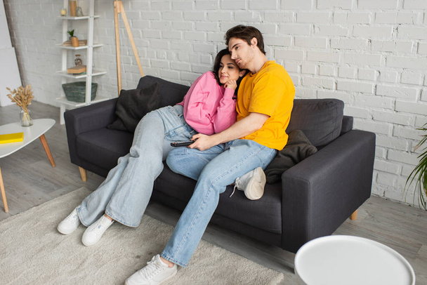 positieve jonge man zitten op de bank, met afstandsbediening en knuffelen vriendin in de woonkamer - Foto, afbeelding