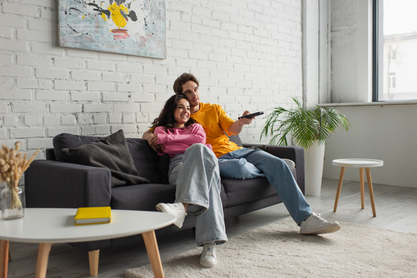 joven positivo sentado en el sofá, sosteniendo el mando a distancia y abrazando a la novia en la sala de estar moderna - Foto, Imagen