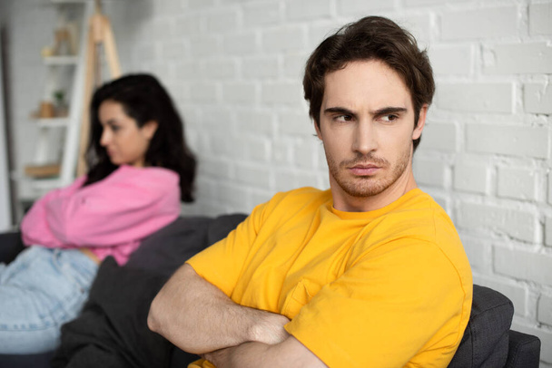 zklamaný mladý muž se zkříženýma rukama sedí na gauči v blízkosti rozmazané přítelkyně doma - Fotografie, Obrázek
