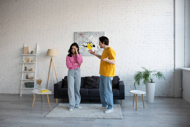 angry young man yelling and gesturing with hands near girlfriend standing in living room - Foto, Imagen