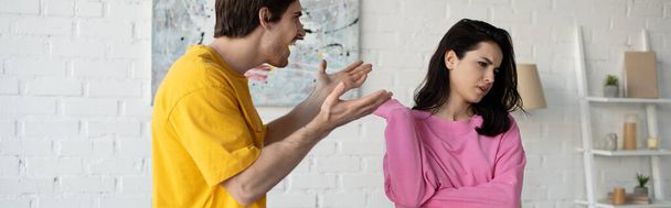 angry young man yelling with outstretched hands near girlfriend in living room, banner - Foto, Bild
