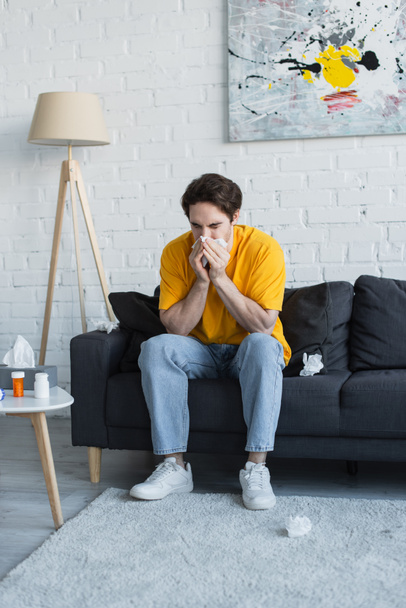 beteg fiatalember ül a kanapén, és fújja orrát papírszalvétával otthon - Fotó, kép