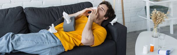 άρρωστος νεαρός άνδρας ξαπλωμένος στον καναπέ και φυσώντας μύτη με χαρτοπετσέτα στο σπίτι, πανό - Φωτογραφία, εικόνα