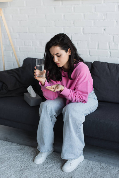 beteg fiatal nő ül a kanapén egy pohár vízzel, és nézi tabletták tenyérben a nappaliban - Fotó, kép
