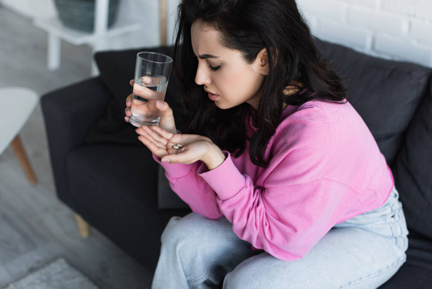 beteg fiatal nő ül a kanapén egy pohár vízzel és tablettákkal a nappaliban - Fotó, kép