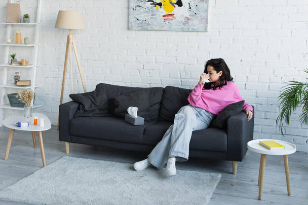 beteg fiatal nő ül a kanapén, és fújja orrát papírszalvétával a nappaliban - Fotó, kép