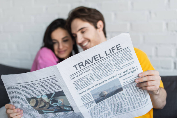 smiling young couple sitting on couch, hugging and holding newspaper at home - Photo, Image