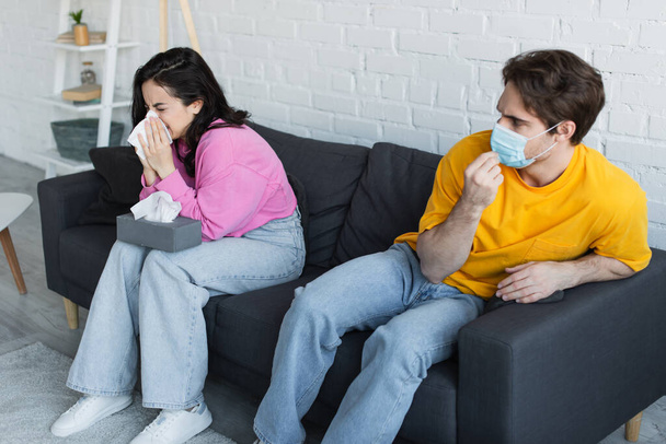 beteg fiatal nő ül a kanapén, és fújja orrát papírszalvétával közel barátja beállítása orvosi maszk otthon - Fotó, kép