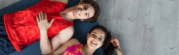 вид сверху на молодую пару в спортивной одежде, лежащую на фитнес-ковриках и смотрящую на камеру дома, баннер - Фото, изображение