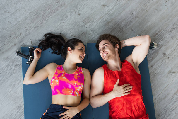 вид сверху на улыбающуюся молодую пару лежащую на фитнес-ковриках рядом с гантелями и смотрящую друг на друга дома - Фото, изображение