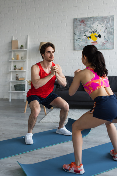 smiling young couple in sportswear doing squats on fitness mats in modern loft - Фото, изображение
