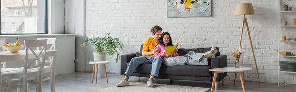 usmívající se mladý muž objímající přítelkyni ležící na gauči s knihou v ruce v obývacím pokoji, prapor - Fotografie, Obrázek