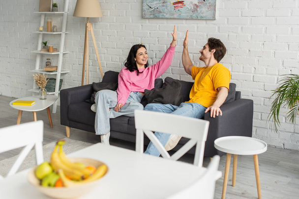 playful young couple sitting on couch with high five gesture in living room - Foto, afbeelding