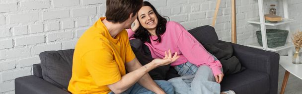 smiling young woman sitting on couch near boyfriend gesturing with outstretched hand in living room, banner - Photo, Image