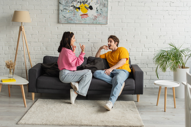 sorrindo jovem casal sentado no sofá e gesticulando com as mãos na sala de estar - Foto, Imagem