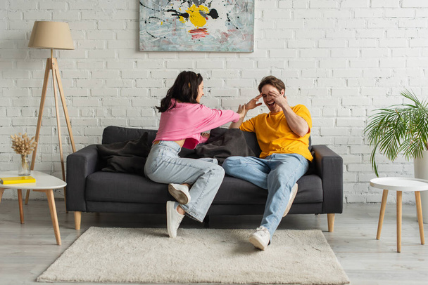 playful young woman touching man covering face with hands on couch in living room - Foto, Imagem