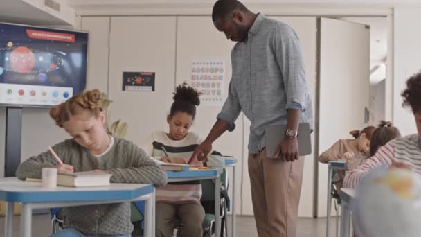 Tilt-up Medium Slowmo junger afroamerikanischer männlicher Lehrer in smarter Freizeitbekleidung, der am Schreibtisch in einem hellen modernen Klassenzimmer steht und Schulmädchen bei der Prüfung in Naturwissenschaften hilft - Filmmaterial, Video