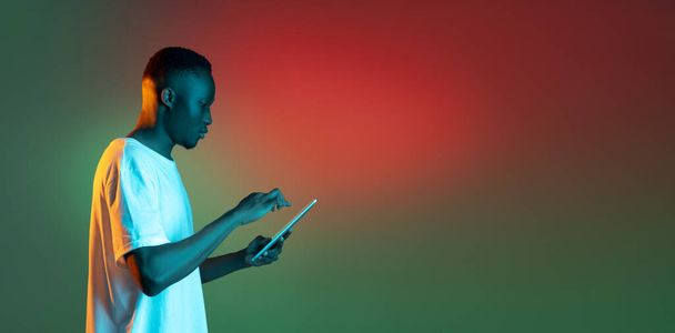 Hombre africano joven usando tableta en luz de neón sobre fondo de gradiente. - Foto, Imagen