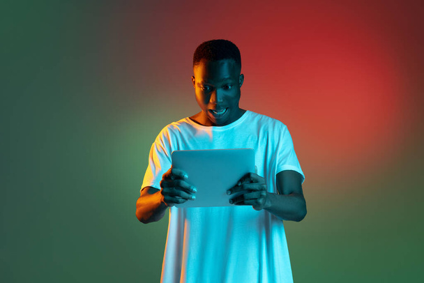 Young african man using tablet in neon light on gradient background. - 写真・画像
