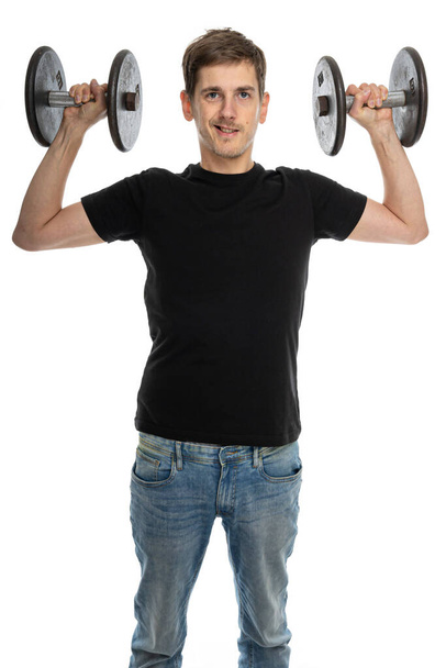 Young handsome tall slim white man with brown hair holds dumbbells looking confident in black shirt in blue jeans isolated on white background - Фото, зображення