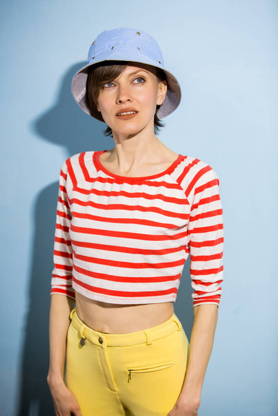 Happy cheerful young woman with short hair, natural makeup over light blue background. Indoor portrait of beautiful brunette young woman wearing blue hat and bright striped clothing - Photo, Image