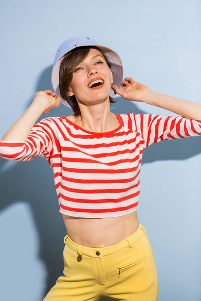Happy cheerful young woman with short hair, natural makeup over light blue background. Indoor portrait of beautiful brunette young woman wearing blue hat and bright striped clothing - Photo, image