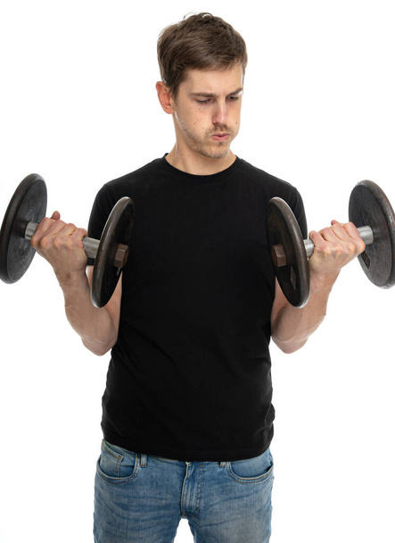Young handsome tall slim white man with brown hair holds dumbbells in black shirt in blue jeans isolated on white background - Valokuva, kuva