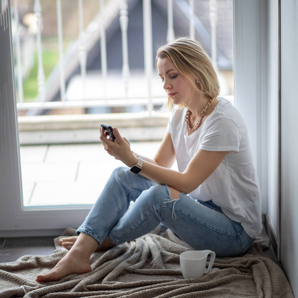 Κορίτσι κάθεται κοντά στο παράθυρο και κρατώντας ένα κινητό τηλέφωνο - Φωτογραφία, εικόνα