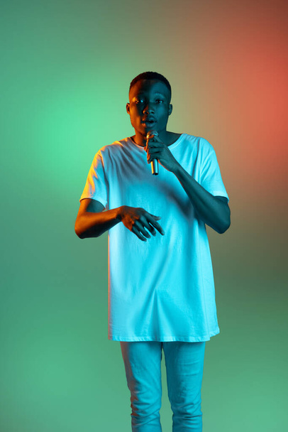 Young african man speaking with microphone on neon gradient background. Concept of emotion, action, facial expression. - Foto, Imagen