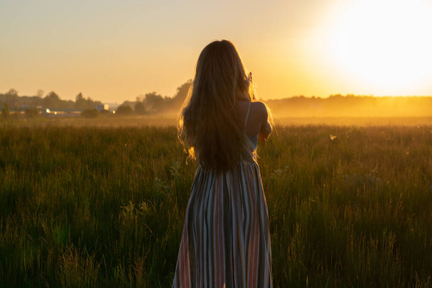 a girl in a dress walks through a field with fog at sunset towards the sun - Photo, Image