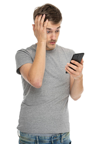 Young handsome tall slim white man with brown hair looks frustrated in gray shirt with mobile phone isolated on white background - Photo, image