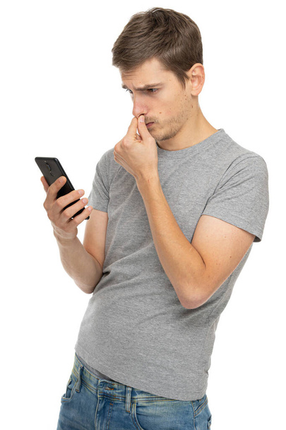 Young handsome tall slim white man with brown hair frowning touching his nose in gray shirt with mobile phone isolated on white background - Fotó, kép