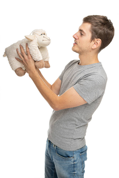 Young handsome tall slim white man with brown hair holding a plush sheep in gray shirt in blue jeans isolated on white background - Foto, Bild