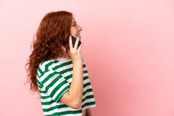 Teenager rothaarige Mädchen über isolierten rosa Hintergrund halten ein Gespräch mit dem Mobiltelefon mit jemandem - Foto, Bild