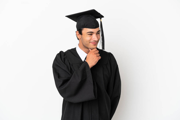 Młody absolwent uniwersytetu ponad odizolowane białe tło patrząc na bok - Zdjęcie, obraz