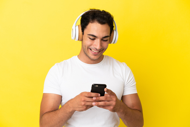 Καυκάσιος όμορφος άντρας πάνω από απομονωμένο κίτρινο φόντο ακούει μουσική και ψάχνει για κινητά - Φωτογραφία, εικόνα