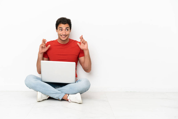 Caucasian handsome man with a laptop sitting on the floor with fingers crossing - Foto, Imagen