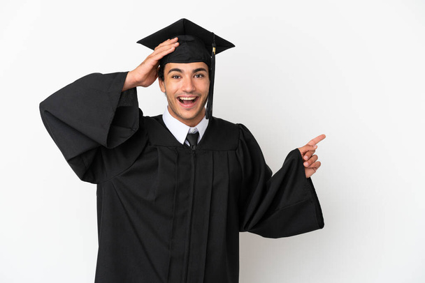 Молодой выпускник университета на изолированном белом фоне удивлен и указывает пальцем в сторону - Фото, изображение