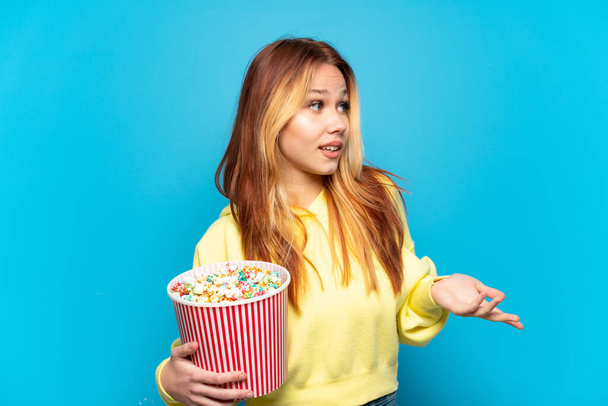 Adolescente chica sosteniendo palomitas de maíz sobre fondo azul aislado con expresión sorpresa mientras mira al lado - Foto, imagen