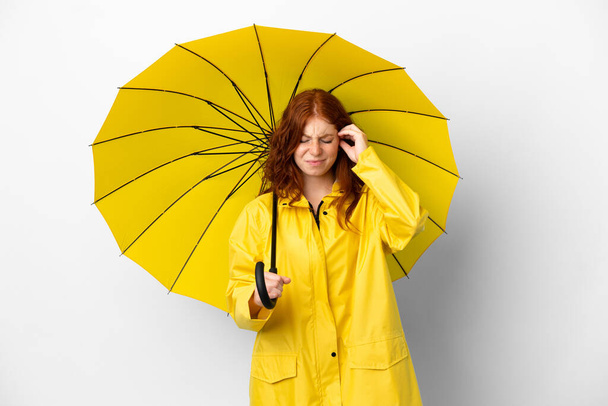 Teenager redhead girl rainproof coat and umbrella isolated on white background frustrated and covering ears - Фото, изображение