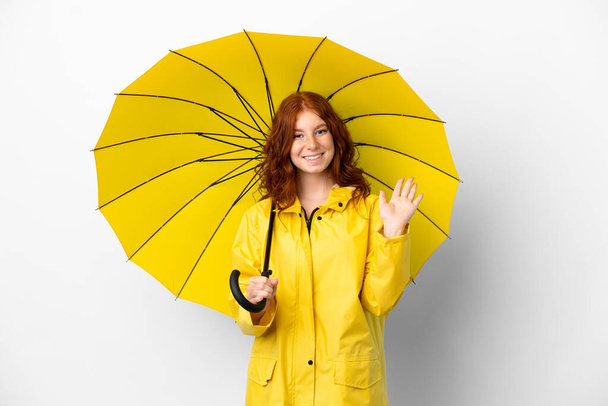 adolescente rossa ragazza impermeabile cappotto e ombrello isolato su sfondo bianco saluto con mano con espressione felice - Foto, immagini