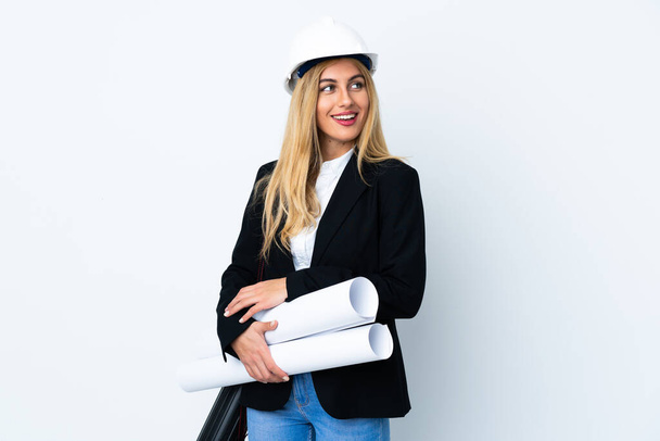 Young architect woman with helmet and holding blueprints over isolated white background looking to the side - Photo, Image