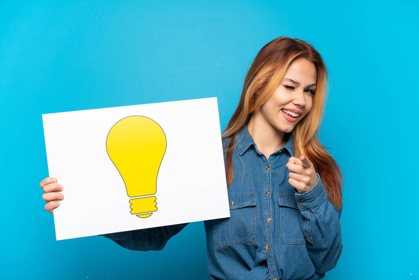 Teenager girl over isolated blue background holding a placard with bulb icon and pointing to the front - Photo, Image