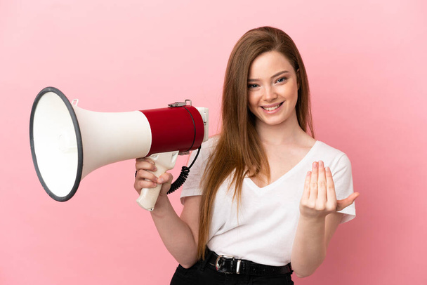 Teenager dívka přes izolované růžové pozadí držící megafon a zvoucí přijít s rukou - Fotografie, Obrázek