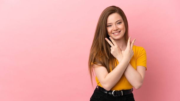 Teenager-Mädchen auf isoliertem rosa Hintergrund lächelt und zeigt Siegeszeichen - Foto, Bild