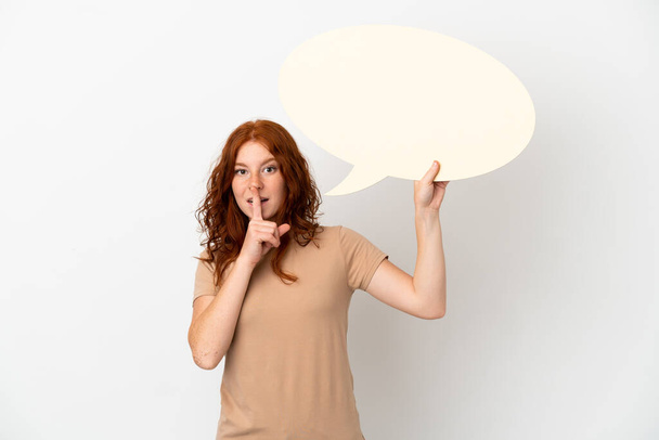 Подростковая рыжая девушка на белом фоне держит пустой пузырь речи и делает жест молчания - Фото, изображение