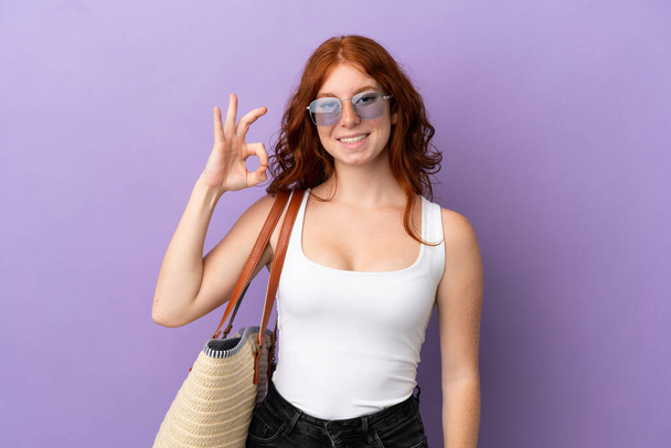 Adolescente pelirroja sosteniendo una bolsa de playa aislada sobre fondo púrpura mostrando signo de ok con los dedos - Foto, Imagen