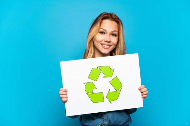 Teenager girl over isolated blue background holding a placard with recycle icon with happy expression - Photo, Image