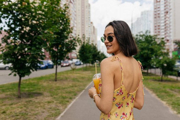 Açık hava portresi, yaz aylarında parlak elbiseler giyen ve gözlüklü şık kısa saçlı bir kadın meyve suyuyla sokakta yürüyor. Yüksek kalite fotoğraf - Fotoğraf, Görsel
