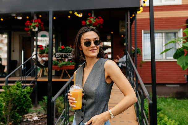 Adorable pretty girl with lovely smile wearing grey vest drinking summer drink and smiling while resting outside. High quality photo - Photo, Image